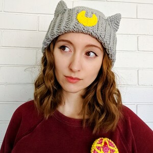 May include: A gray knitted beanie with cat ears and a yellow crescent moon appliqu&eacute;. The beanie is worn by a person with long brown hair and a red sweater. The person is wearing a pink and yellow crocheted flower pin.