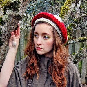 Peut inclure: Une femme portant un chapeau en crochet en forme de champignon rouge et blanc. Le chapeau a un dessus blanc et un dessous rouge avec des rayures blanches. La femme a de longs cheveux bruns ondul&eacute;s et porte une veste &agrave; capuche marron.