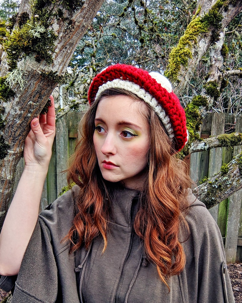 Peut inclure: Une femme portant un chapeau en crochet en forme de champignon rouge et blanc. Le chapeau a un dessus blanc et un dessous rouge avec des rayures blanches. La femme a de longs cheveux bruns ondul&eacute;s et porte une veste &agrave; capuche marron.
