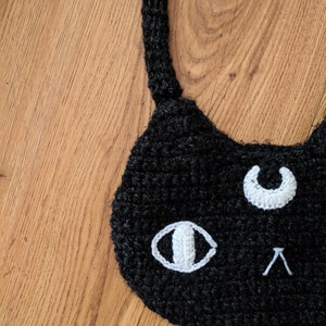 May include: Black crocheted cat face with white crocheted eyes and a crescent moon. The cat face is on a wooden surface.