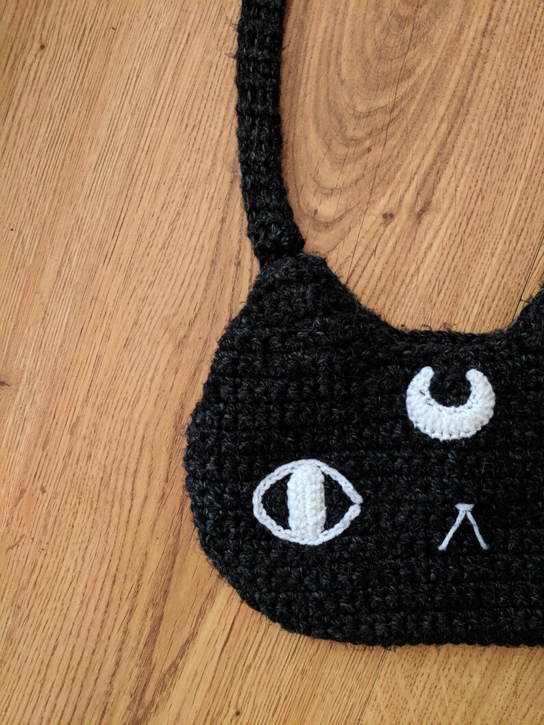 May include: Black crocheted cat face with white crocheted eyes and a crescent moon. The cat face is on a wooden surface.