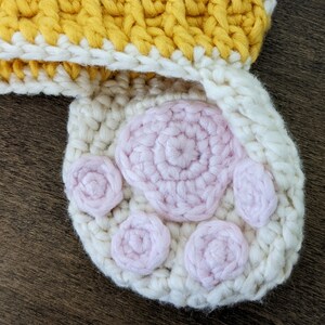 May include: A white crocheted hat with a yellow and white striped band and a pink crocheted paw print design.