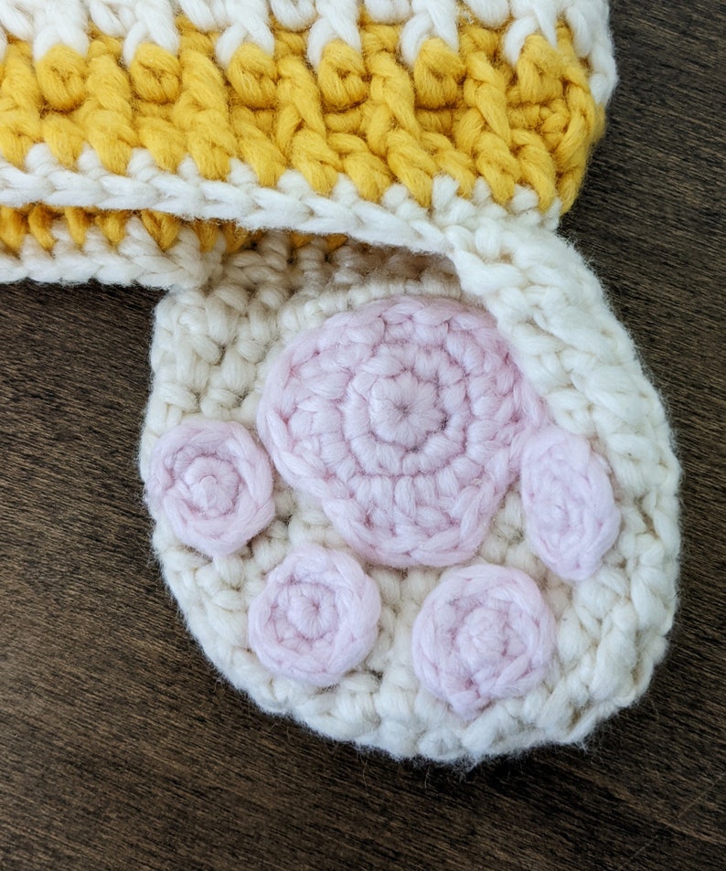 May include: A white crocheted hat with a yellow and white striped band and a pink crocheted paw print design.