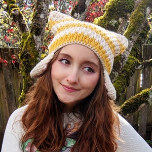 May include: A woman wearing a yellow and white striped crocheted hat with cat ears. The hat has a pointed top and is made with a soft yarn.