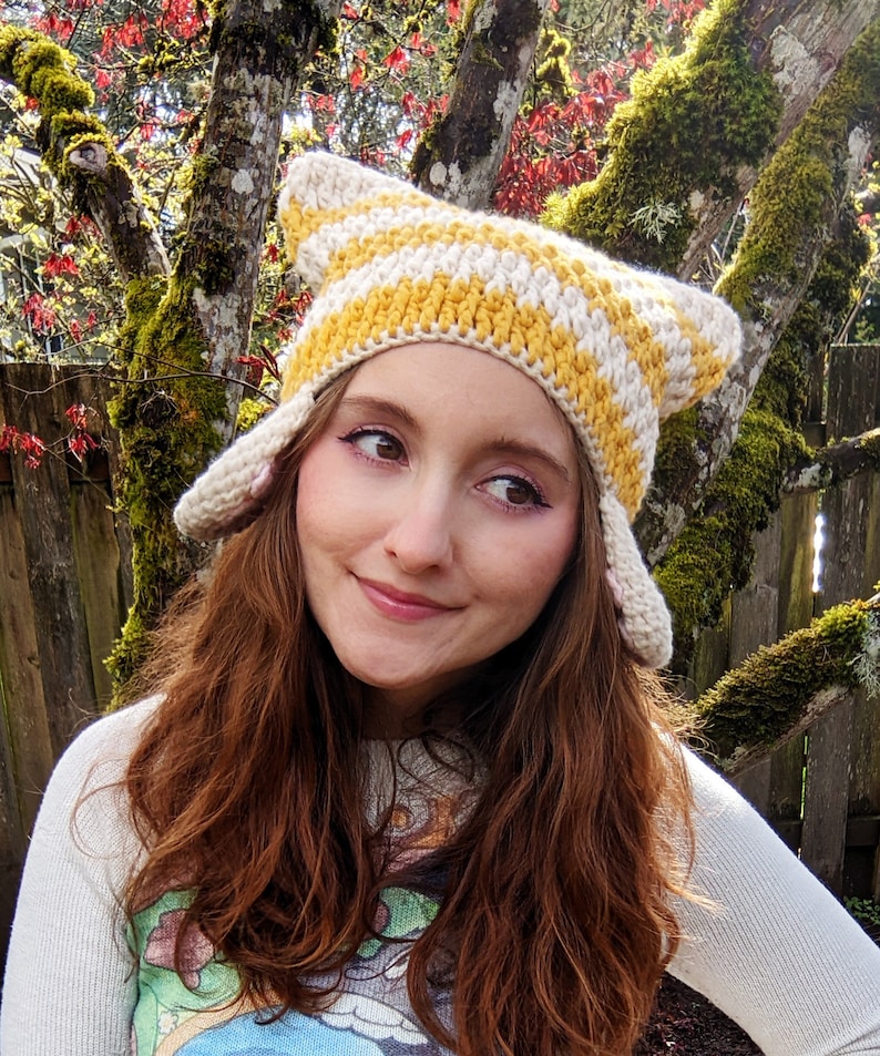 May include: A woman wearing a yellow and white striped crocheted hat with cat ears. The hat has a pointed top and is made with a soft yarn.