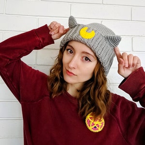 May include: A woman wearing a gray crocheted beanie with cat ears and a yellow crescent moon design. She is also wearing a maroon sweater and a crocheted pin with a yellow and pink design.