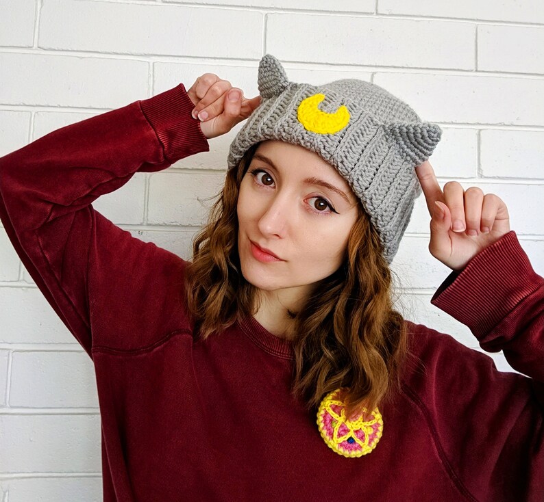 May include: A woman wearing a gray crocheted beanie with cat ears and a yellow crescent moon design. She is also wearing a maroon sweater and a crocheted pin with a yellow and pink design.
