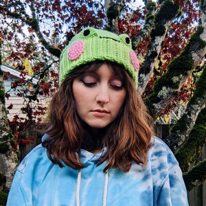 May include: A person wearing a green crocheted frog hat with pink cheeks. The hat is worn over brown hair and the person is wearing a blue tie-dye hoodie.