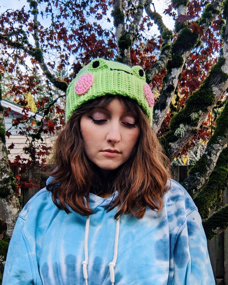 May include: A person wearing a green crocheted frog hat with pink cheeks. The hat is worn over brown hair and the person is wearing a blue tie-dye hoodie.