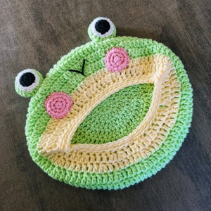 May include: A handmade, crocheted frog-shaped item in shades of green, yellow, pink, and black. The frog has large, round eyes, pink cheeks, and a smiling mouth. The item is circular with a central opening.