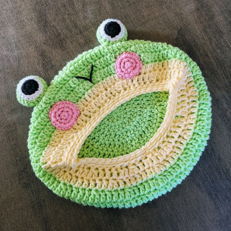 May include: A handmade, crocheted frog-shaped item in shades of green, yellow, pink, and black. The frog has large, round eyes, pink cheeks, and a smiling mouth. The item is circular with a central opening.