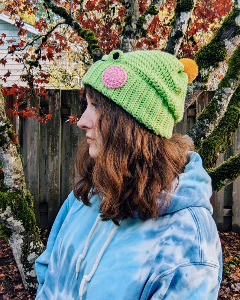 May include: A person wearing a green crocheted hat with a pink and yellow frog design. The hat has a pom-pom on top. The person is wearing a blue tie-dye hoodie.