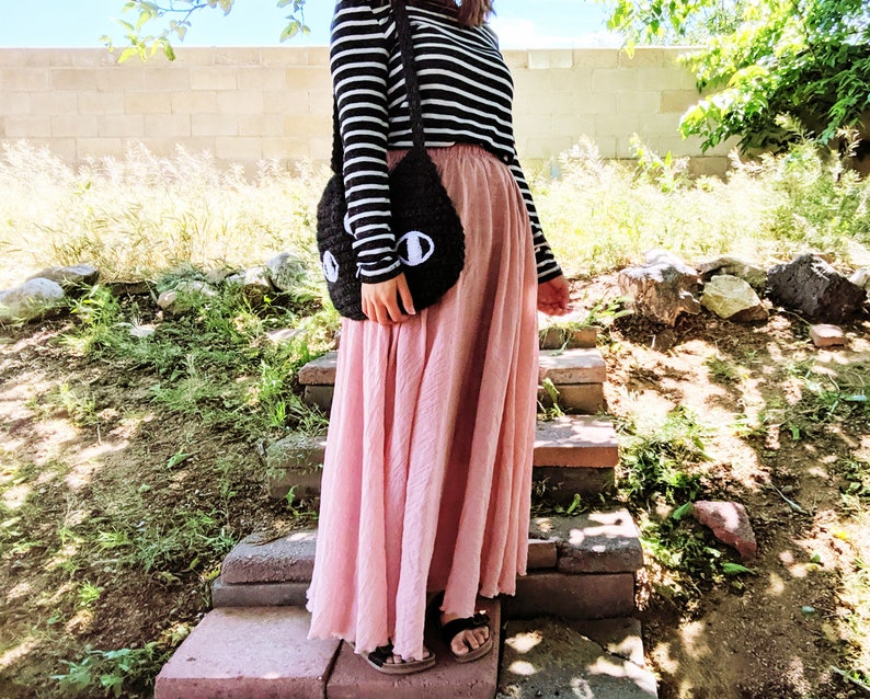 May include: A woman wearing a black and white striped long-sleeved shirt and a long pink skirt. She is carrying a black crocheted bag with white eyes.