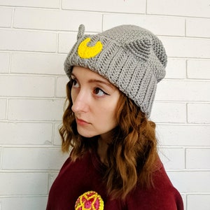 May include: Gray crocheted beanie with a yellow crescent moon and cat ears. The beanie is worn by a person with long brown hair.