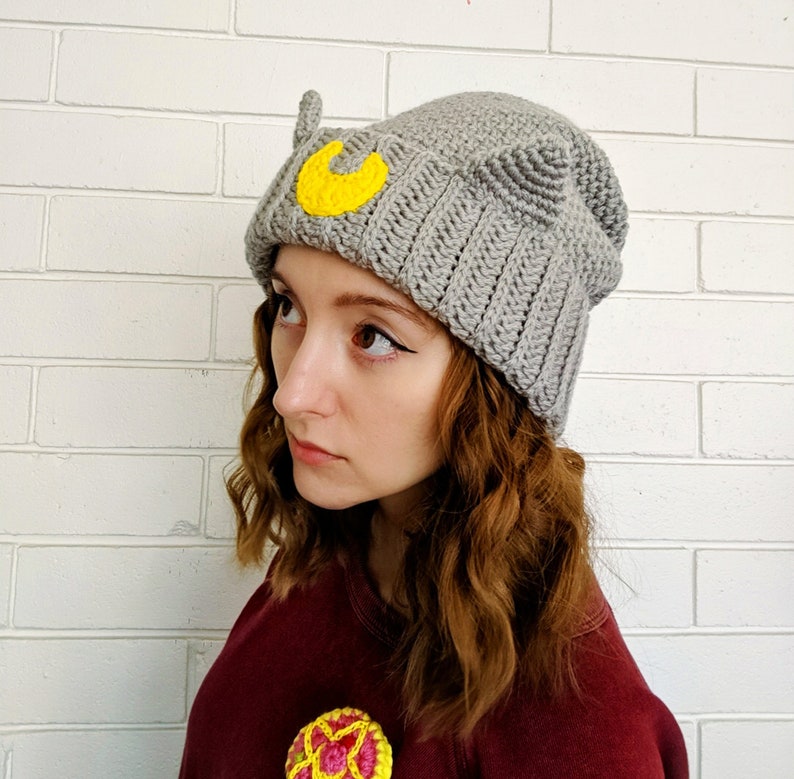 May include: Gray crocheted beanie with a yellow crescent moon and cat ears. The beanie is worn by a person with long brown hair.