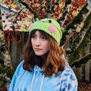May include: A person wearing a green crocheted frog hat with pink accents. The hat has a textured surface and is worn over brown hair. The person is wearing a blue tie-dye hoodie.