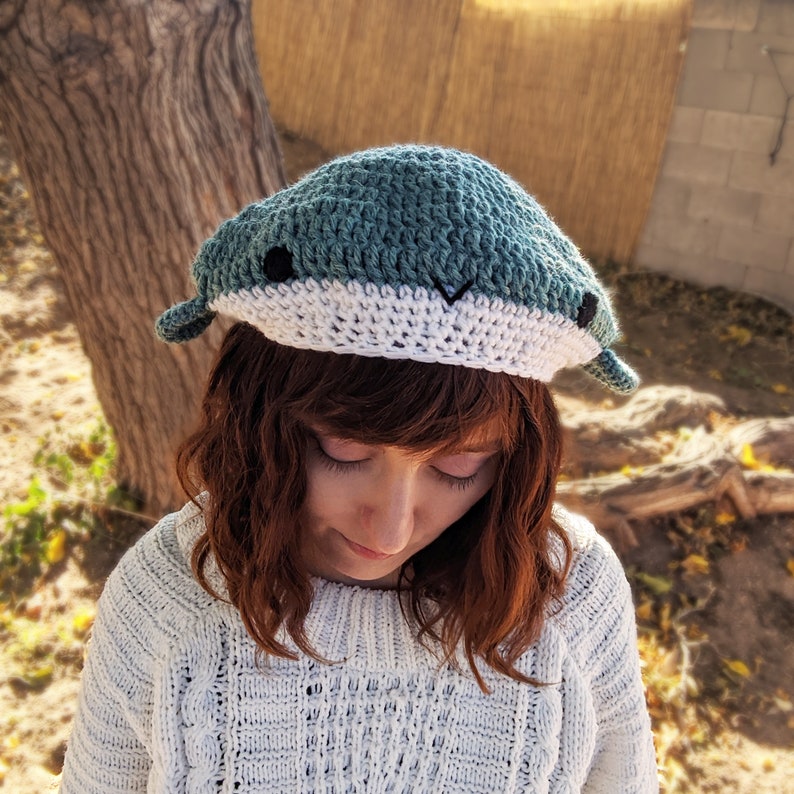 Pu&ograve; includere: Una persona indossa un cappello di balena all'uncinetto blu e bianco. Il cappello ha occhi neri e un sorriso bianco.