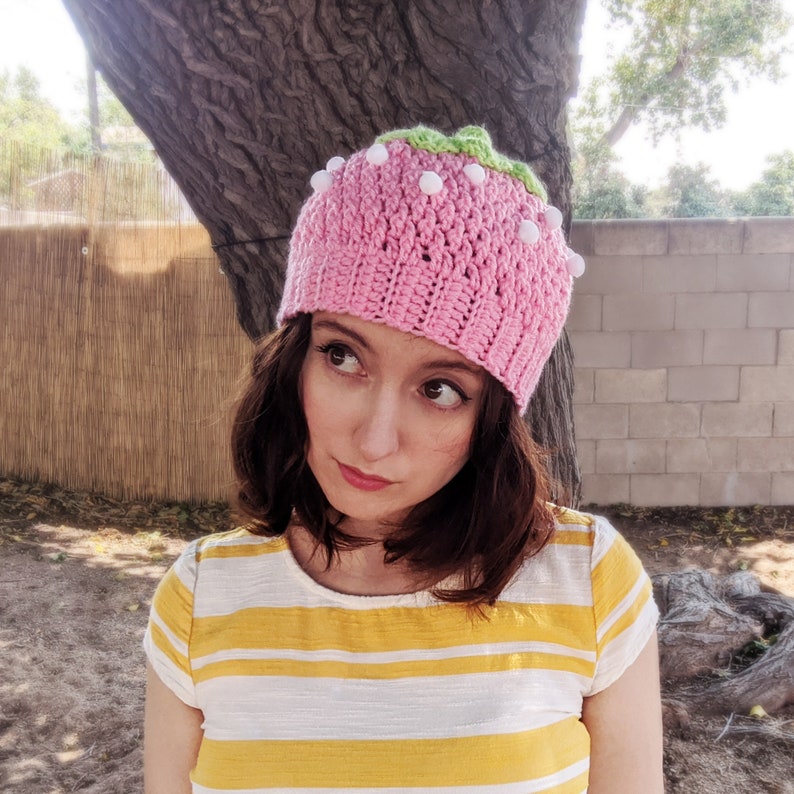 May include: A pink crocheted hat shaped like a strawberry with white pom-poms. The hat is worn by a person with brown hair.