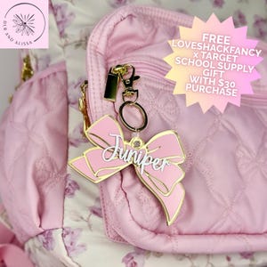 May include: A light pink bow-shaped keychain with gold-tone metal edges and the name Juniper written in white script lettering. The keychain is attached to a gold-tone metal lobster clasp and ring, resting against a quilted light pink backpack.