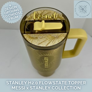May include: A gold-coloured Stanley H2.0 FlowState tumbler featuring a matching gold handle and a lid with a yellow straw cover. The lid includes a gold glitter sunburst design and a custom gold name plate reading Leonel.
