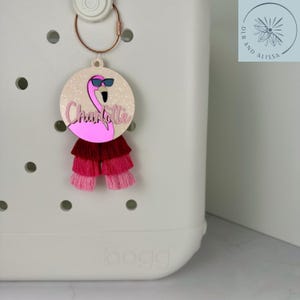 May include: A pink flamingo charm with sunglasses and the name "Charlotte" in cursive. The charm has a layered tassel in shades of pink and red. Attached to a rose gold keyring.