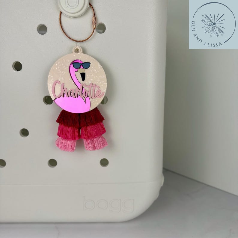 May include: A pink flamingo charm with sunglasses and the name "Charlotte" in cursive. The charm has a layered tassel in shades of pink and red. Attached to a rose gold keyring.