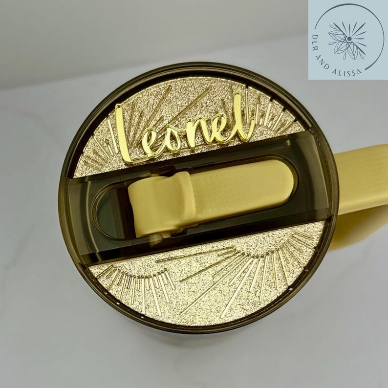 May include: A circular dark-tinted tumbler lid featuring a central yellow flip-top straw opening. The lid surface is decorated with a gold-coloured textured sunburst pattern and the name Leonel in a gold-coloured cursive script font.