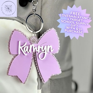 May include: A lavender bow-shaped keychain featuring a silver metal ring and a white script name tag that reads Kamryn. The edges of the bow are lined with small, sparkling lavender crystals, and the entire piece is attached to a grey backpack.