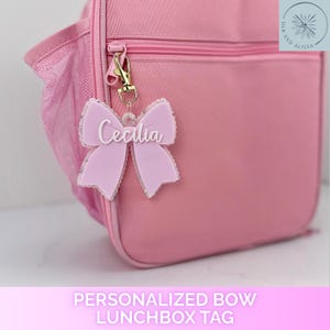 May include: A light pink lunchbox with a matching light pink acrylic bow-shaped bag tag attached to the zipper. The tag features the name Cecilia in white script lettering and is secured with a gold-toned metal lobster clasp.