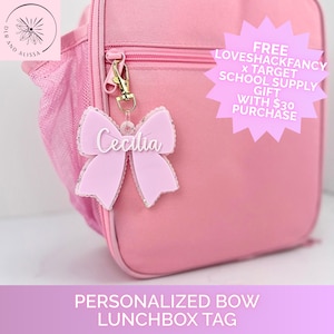 May include: A light pink acrylic bow-shaped bag tag featuring the name Cecilia in white script lettering. The tag is attached to a pink lunchbox with a gold-toned metal lobster clasp. The text on the image reads FREE LOVESHACKFANCY X TARGET SCHOOL SUPPLY GIFT WITH $30 PURCHASE.
