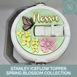 May include: A white Stanley Iceflow topper with a light green background, featuring a gold "Nessa" name, a brown butterfly, yellow and pink flower accents. The text "STANLEY ICEFLOW TOPPER SPRING BLOSSOM COLLECTION" is at the bottom.