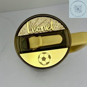 May include: A dark-tinted tumbler lid featuring a gold-coloured sliding closure and a gold-coloured accent plate with a black football graphic. The lid includes a gold-coloured nameplate with the name Leonel in a cursive script font.