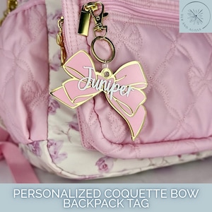 May include: A light pink acrylic bow-shaped bag tag featuring the name Juniper in white script lettering. The tag is attached to a quilted light pink backpack with a gold-toned metal lobster clasp and a circular ring, creating a decorative accessory.