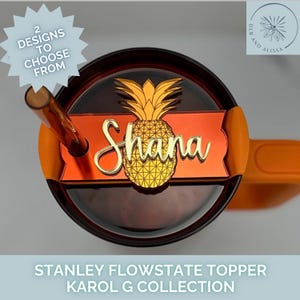 May include: A Stanley Flowstate topper with a pineapple design and the name "Shana" in gold script. The topper is orange and gold, with the text "2 Designs to Choose From". The product is from the Karol G Collection.