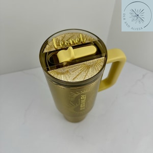 May include: A gold-coloured tumbler with a handle and a clear lid featuring a gold glitter insert. The lid has a light yellow sliding closure and a gold name tag that reads Leonel. The side of the tumbler displays the Stanley logo in gold.