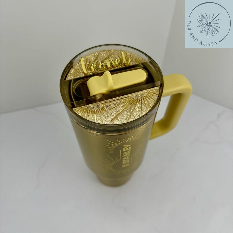 May include: A gold-coloured tumbler with a handle and a clear lid featuring a gold glitter insert. The lid has a light yellow sliding closure and a gold name tag that reads Leonel. The side of the tumbler displays the Stanley logo in gold.