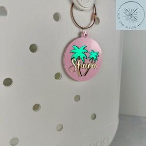 May include: A pink, round bag tag with a gold-colored wire ring. The tag features a green palm tree design and the name "Shana" in gold lettering. The tag is attached to a white bag.