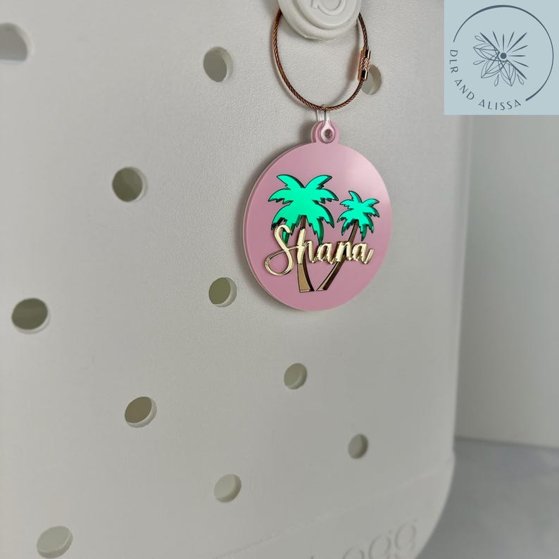 May include: A pink, round bag tag with a gold-colored wire ring. The tag features a green palm tree design and the name "Shana" in gold lettering. The tag is attached to a white bag.