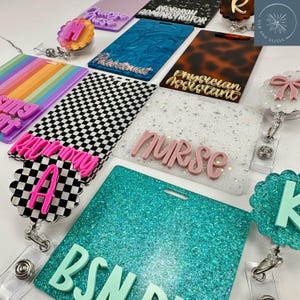May include: A collection of rectangular acrylic badge reels and ID holders featuring various designs including rainbow stripes, black and white checkers, glitter finishes, and tortoise patterns, with raised lettering displaying titles like Nurse, BSN, and Phlebotomist.