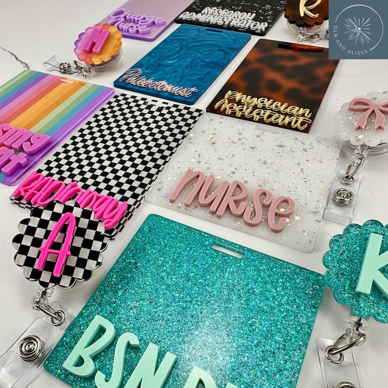 May include: A collection of rectangular acrylic badge reels and ID holders featuring various designs including rainbow stripes, black and white checkers, glitter finishes, and tortoise patterns, with raised lettering displaying titles like Nurse, BSN, and Phlebotomist.
