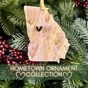 May include: A Georgia-shaped ornament with a heart-shaped accent. The ornament has a pink and gold marbled design and is suspended by a gold string. The text "Hometown Ornament Collection" is displayed below the ornament.