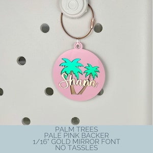 May include: A round, pale pink charm with a gold-colored wire and clasp. The charm features two green palm trees and the name "Shana" in gold script. The charm is attached to a white object.