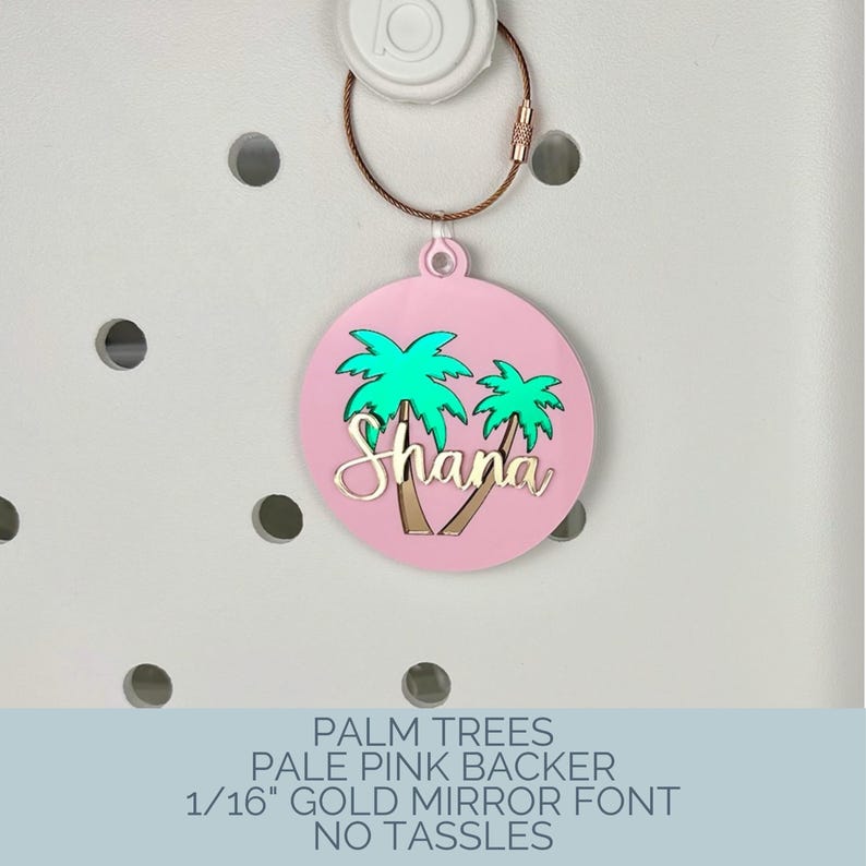 May include: A round, pale pink charm with a gold-colored wire and clasp. The charm features two green palm trees and the name "Shana" in gold script. The charm is attached to a white object.