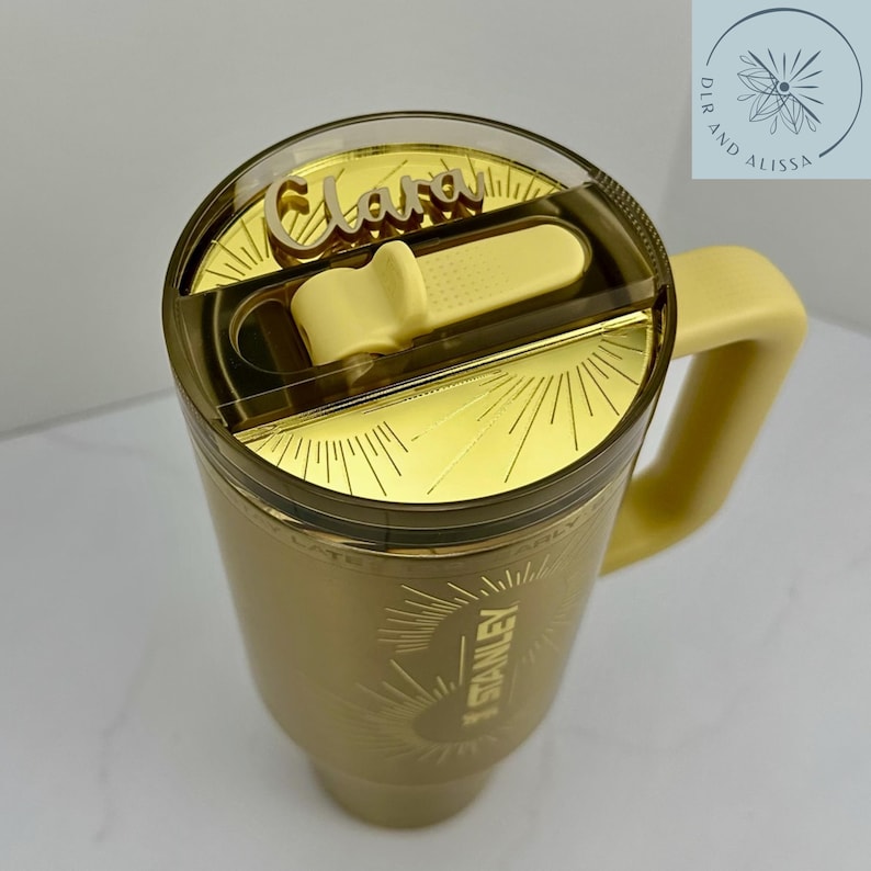 May include: A gold-coloured tumbler with a matching handle and a clear lid featuring a gold sunburst design. The lid includes a yellow sliding closure and a custom name tag that reads Clara in a script font. The word Stanley is visible on the side.