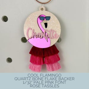 May include: A round, pink flamingo charm with the name "Charlotte" in cursive. The flamingo wears sunglasses and is attached to a layered tassel in shades of pink and red. The charm has a bone flake backer.