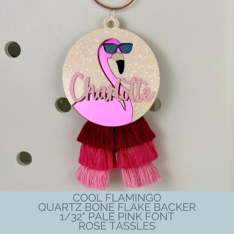 May include: A round, pink flamingo charm with the name "Charlotte" in cursive. The flamingo wears sunglasses and is attached to a layered tassel in shades of pink and red. The charm has a bone flake backer.