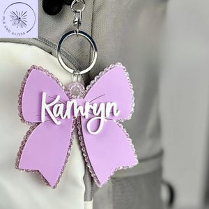 May include: A lavender bow-shaped keychain featuring a silver metal ring and clasp. The bow has a glittery border and the white script name Kamryn is attached to the center. The design includes a small circular accent above the name.