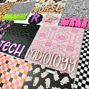 May include: A collection of rectangular acrylic badge reels featuring various patterns including pink pumpkins with bows, black bats, ghosts, and checkered prints. Several pieces feature raised, colorful acrylic lettering spelling out titles like RADIOLOGY, TECH, NURSE, and CNA.