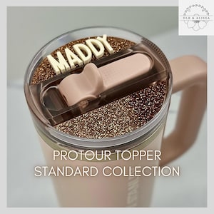 May include: A pink travel mug with a clear lid, featuring the name "MADDY" in white letters. The lid has a glittery brown accent and a pink sliding closure. The mug has the text "PROTOUR TOPPER STANDARD COLLECTION".