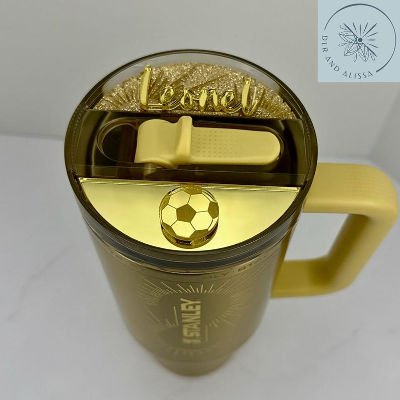 May include: A gold-coloured Stanley tumbler with a handle, featuring a clear lid with a yellow sliding closure. The lid is decorated with a gold football charm and a gold script nameplate reading Leonel over a gold glitter background.